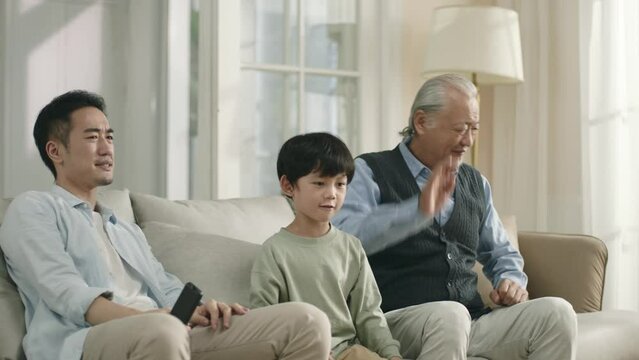 asian grandfather father son sitting on couch at home watching soccer game on tv and disappointed at team's performance