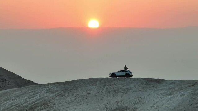 Man sits on a desert hill overlooking a sunrise in ontop of a Mazda car; Israel in the Judean Desert looking out to Jordan. 