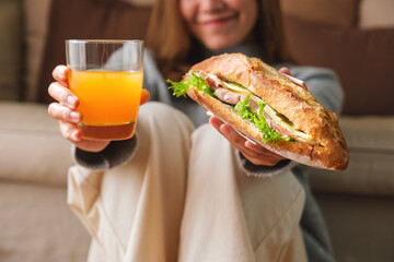 Closeup image of a young woman holding and eating french baguette sandwich and orange juice at home - Powered by Adobe