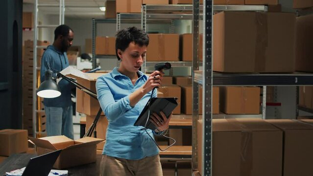 Female employee using scanner and tablet to do stock inventory, scanning bar code from packages on storehouse racks. Woman checking merchandise in boxes, supply chain logistics.
