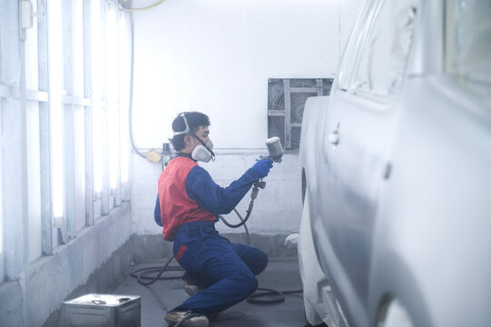 Asian auto mechanic wearing protective suit and respirator paints car body bumpers in paint room,spraying cars in paint room