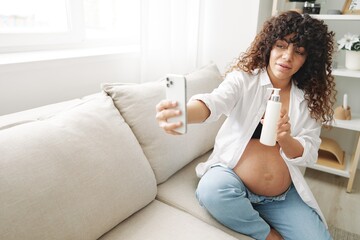 A pregnant woman blogger advertises a cream for stretch marks on the body during pregnancy, filming herself on the phone while sitting on the couch at home