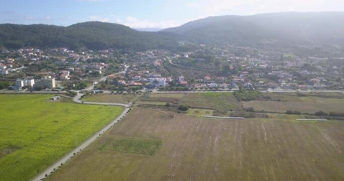 A small town with the same brown houses and roofs, through which a wide asphalt road passes, is located between green fields and mountains, near some houses there is a pool with clear blue water