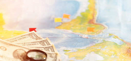 Pile of US dollar cash, magnifying glass and pushpin - travel concept. Destination marked with a pin on the map.