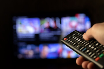 A person holds a remote control in his hand and watches TV with a blurry TV screen in the...