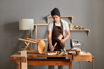 Portrait of man carpenter wearing brown apron and black cap using mallet and chisel in his workshop...