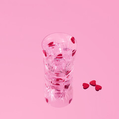 Crystal drinking glass with ice cubes and red hearts with mirror reflection. Creative Valentines day celebrating concept. Minimal nostalgic love aesthetic. Retro pink background.