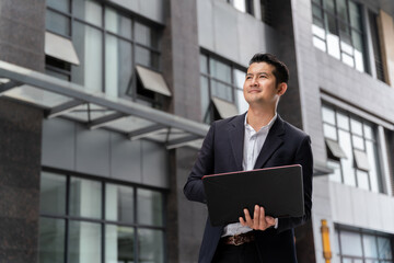 Handsome Asian business man holding laptop looking away happily in urban public space.