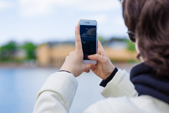 Young woman on vacation using mobile phone to take pictures river and city, wearing a white coat and sunglasses. Rest and relaxation, lifestyle concept.