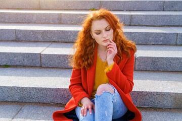 Outdoors fashion portrait young elegant woman in red coat and yellow sweater, curly red hair develops in the wind, in the park on a step