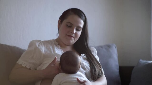 Mam&aacute; latina sentada en sof&aacute; amantando a bebe mexicano en posici&oacute;n de koala, ni&ntilde;a durmiendo.
