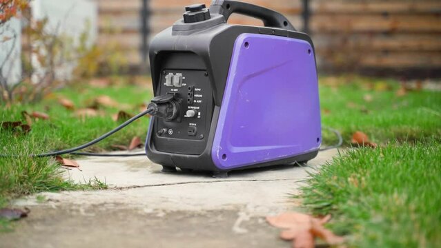A small gasoline inverter generator with a wire connected to a socket works on the street close-up. Purple power generator with a plastic case. Alternative power source in case of blackout