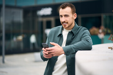 Smiling handsome man holding smartphone using mobile app shopping online standing on the street. Modern hipster chatting looking at camera. Mobile banking concept 