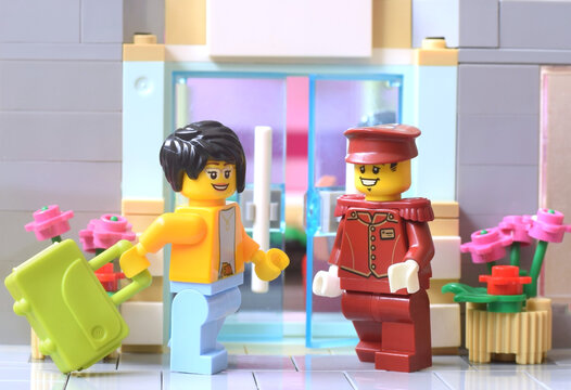 Lego minifigure of girl is arriving in a hotel with a doorman in uniform. Editorial illustrative image of popular plastic bricks toy. Studio shot.