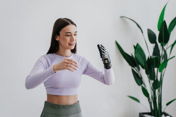 Beautiful European brunette girl in sportswear adjusting hi tech arm prosthesis at home against large plant on background. Orthopaedics and medicine for people after trauma. Hi tech medicine.
