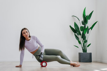 Gorgeous brunette European girl in sportswear makes exercise at home adjusts massaging roll by artificial bionic arm, looks at camera, against minimalistic interior with big plant. Orthopaedics