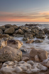 Ocean scene with low sun on the horizon, some rocks in foreground and mid ground with blurred waves around them plus a few colourful clouds.