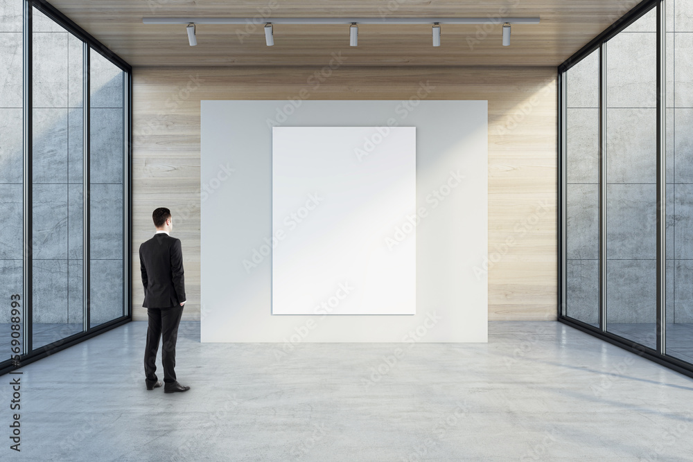 Poster Man in black suit back view looking at blank white poster with space for your logo or text on light partition on light wooden background in empty exhibition hall with transparent walls, mock up