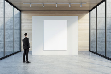 Man in black suit back view looking at blank white poster with space for your logo or text on light partition on light wooden background in empty exhibition hall with transparent walls, mock up