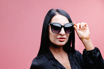 Young caucasian girl looking at camera through sun glasses on pink background. Dark-haired beauty wears black shirt. People sincere emotions lifestyle concept.
