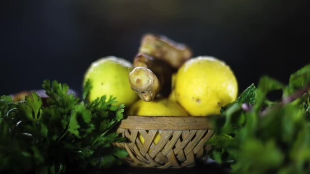 Cinematic close-up shot of fresh lemons, ginger coriander, and some mint leaves on a black colored shiny wooden surface. Fresh green vegetables in wicker basket.