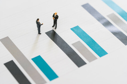 Two Miniature Businessmen Speak And Handshake Stand On The Bar Chart, As Commitment, Partnership, Business And Investment Concept.