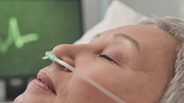 Closeup of mature Asian woman with nasal cannula lying in hospital bed, rehabilitating after surgery operation