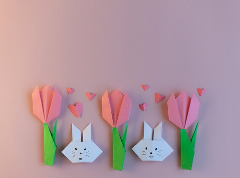 Easter paper crafts for children. Flowers tulips and white rabbits made of origami paper on a pastel pink background. Spring decor, decoration for children, greeting card