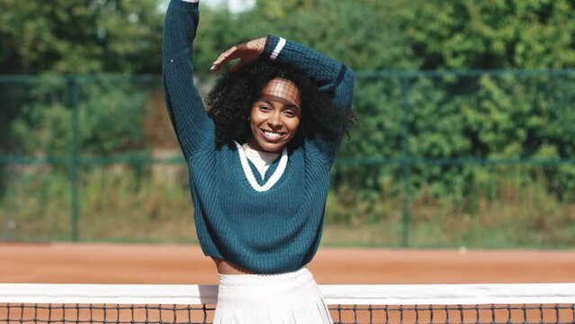 4k video. Young beautiful african american woman posing on clay tennis court with retro vintage tennis rocket. Sunny day outdoor. Sport, 80s, wellness concept.