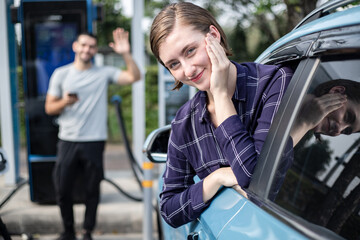 Fototapeta na wymiar Young beautiful woman and man traveling by electric car having stop at charging station standing leaning on window vehicle with bright smile.