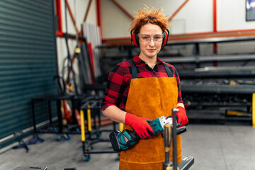 A young girl with curly red hair who is an apprentice in a metal workshop is using tools, she is wearing protective equipment