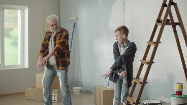Grandson is teaching grandpa how to dance and showing modern dance moves with his hands. Elderly man and young guy are dancing merrily and smiling joyfully.
