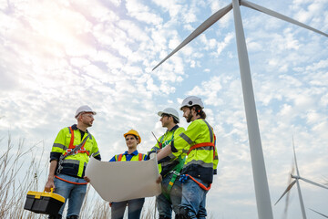 Teamwork engineer worker wearing safety uniform holding and reading blueprint at wind turbine field...