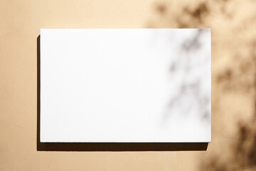 White canvas, blank picture mockup hanging on beige wall with dark shadows of leaves. Poster...