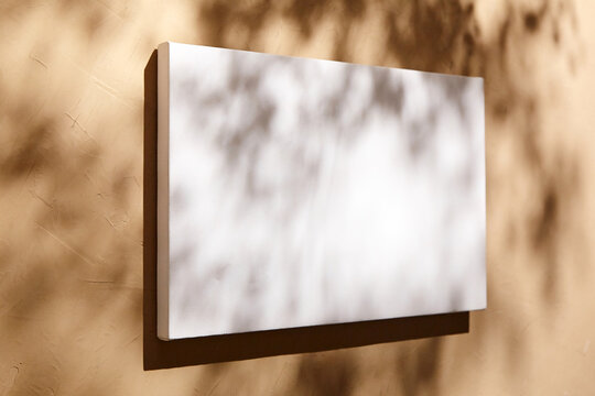 White canvas, blank picture mockup hanging on beige wall with dark shadows of leaves. Poster mockup, empty canvas with shadows of plant, side view