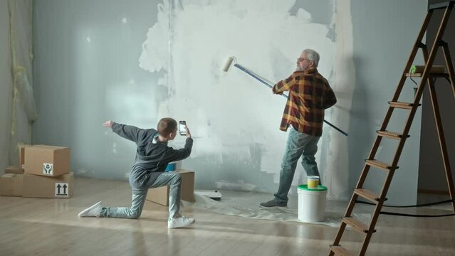 Grandson taking photo with mobile phone and filming a video of grandpa painting a wall white with a paint roller. Elderly man doing repairs in apartment and posing for photo for memory.
