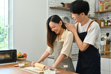 Fototapeta premium Romantic young couple preparing homemade pastry, kneading dough with a rolling pin on wooden table in kitchen