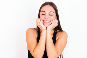 Fototapeta premium Portrait of young woman wearing sportswear over white studio background being overwhelmed, expressing excitement and happiness with closed eyes and hands near face.
