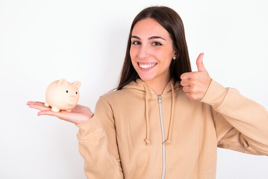 Young Woman Wearing Beige Sweater Over White Studio Background Holding Piggy Bank And Rising Thumb Up Making Approval Sign. Savings And Economy Concept. 