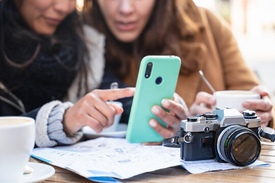 Woman friends looking at cell phone sitting in cafe with photo camera, checking tourist city map