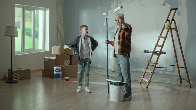 Grandpa planning repairs in apartment and explaining to his grandson the amount of work. Eelderly man with roller in hands talking about work, while teenager hides behind stack of cardboard boxes.