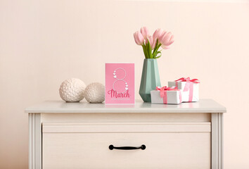 Vase with tulips, gifts and greeting card for Women's Day on chest of drawers near light wall