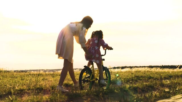 mother teaches child to ride a bicycle at sunset. happy family concept. child girl pedaling a two-wheeled bicycle sun. vacation kid with parent nature. child daughter rides bicycle. happy family