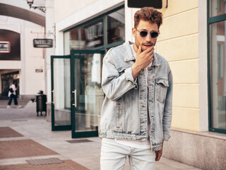 Portrait of handsome confident stylish hipster model. Sexy man dressed in jeans jacket. Fashion male posing in the street. In sunglasses in Europe city. Outdoors at sunset