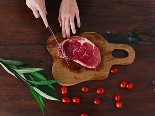 Woman with knife in hand cutting fresh steak meat for roasting in kitchen with salt pepper and other spices on table, red cherry tomatoes and herbs, preparing dinner. wooden table, top view.
