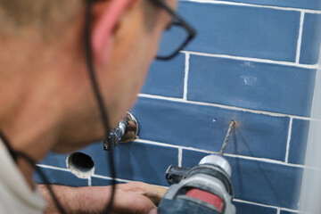 Unrecognizable senior mason drilling of a hole in the wall of blue tiles.