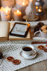 Fototapeta na wymiar Cup of tea, books, tablet, bowl of cookies, various spices, pine cones and lit candles. Hygge at home. Selective focus.