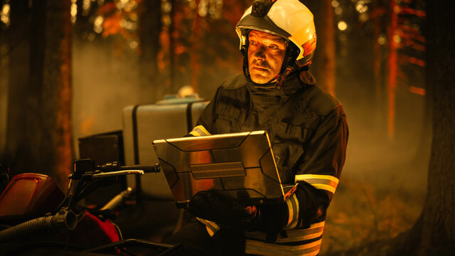Portrait of a Handsome Fireman in Safety Gear Using Heavy-Duty Laptop Computer, Reporting on a Situation with a Dangerous Wildland Fire in a Forest. Hard Day at Work for a Firefighter Volunteer.