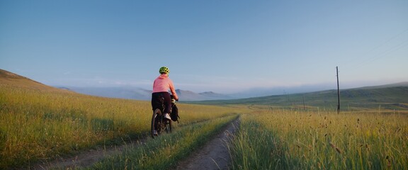 The athlete trains for a long distance. Rear view. The picturesque landscape with a field in the fog. The woman on bike with bikepacking bags in activewear in green black colors