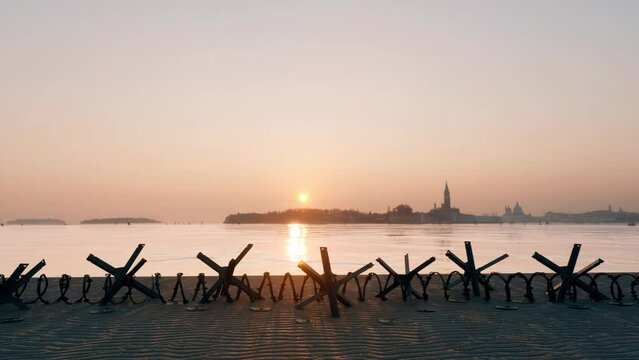 A city shoreline at sunset, dotted with strategic barriers including landmines, barbed wire, and metal anti-tank hedgehogs.